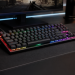 Teclado mecânico gamer com iluminação RGB sobre uma mesa, ao lado de um mouse e monitor